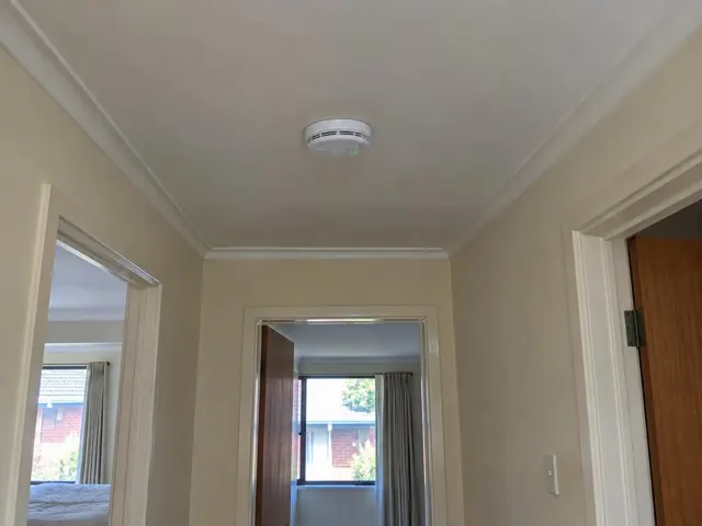 Ceiling-mounted smoke alarm in a cream hallway with doorways and crown moulding in Matraville